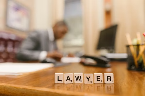Scrabble tiles spelling "LAWYER" on a desk with an attorney working in the background.